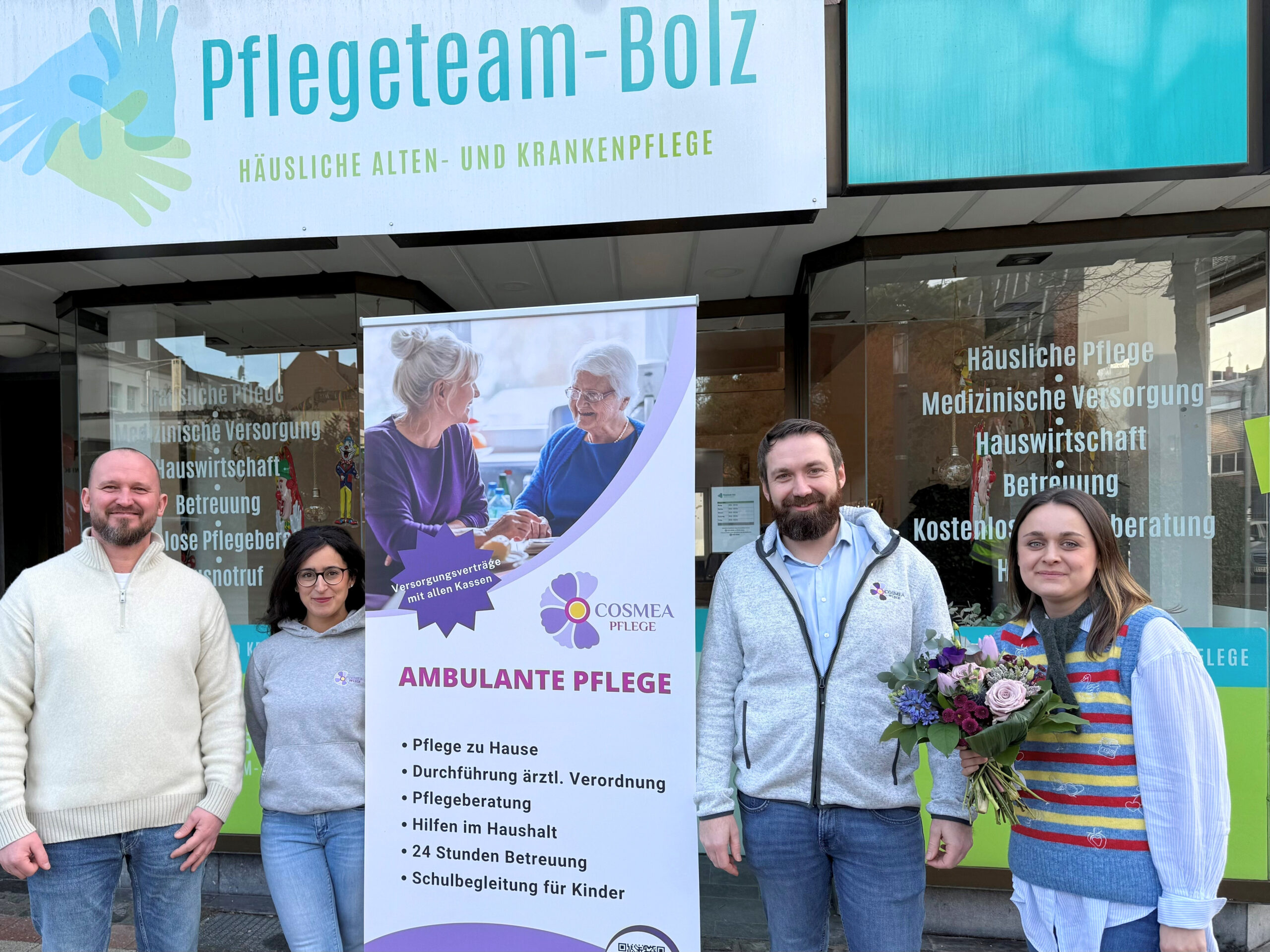 Vier Mitarbeitende eines ambulanten Pflegedienstes stehen vor einem Ladenlokal mit der Aufschrift „Pflegeteam Bolz“. In der Mitte befindet sich ein Roll-up-Banner von Cosmea Pflege mit Informationen zur ambulanten Pflege. Eine Person hält einen Blumenstrauß, alle blicken freundlich in die Kamera.